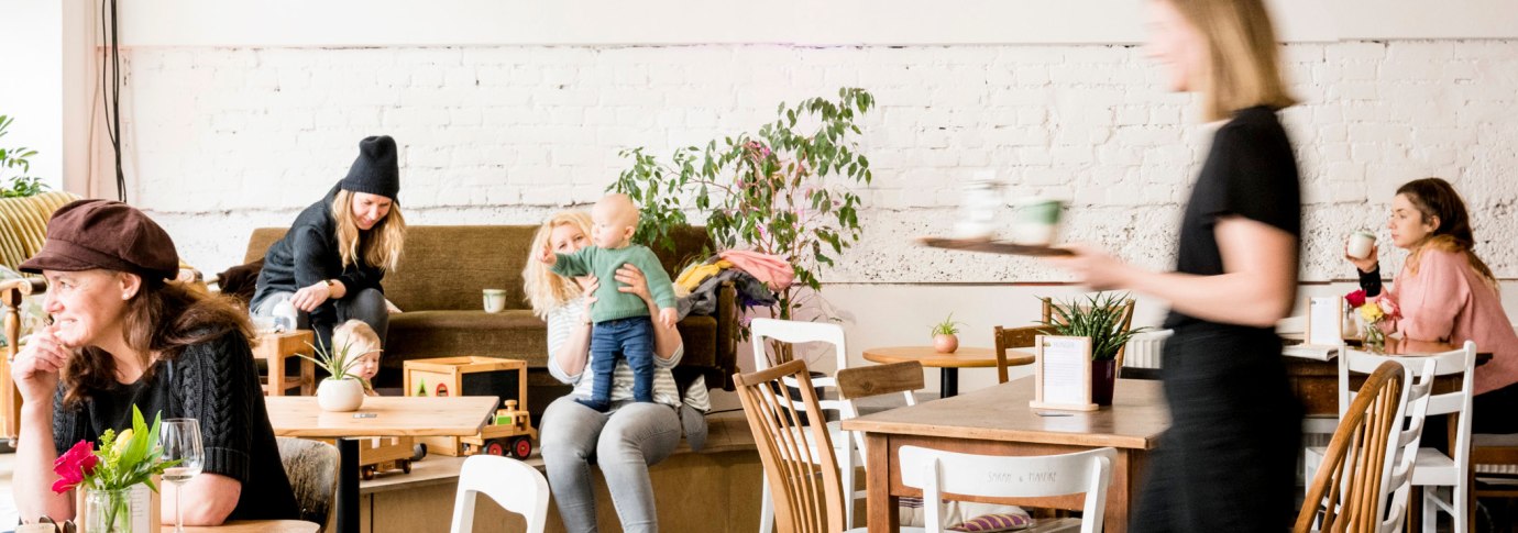 Gem&uuml;tliches Caf&eacute; mit spielenden Kindern und Erwachsenen. Eine Bedienung serviert Getr&auml;nke. Pflanzen und Holzm&ouml;bel schaffen eine einladende Atmosph&auml;re., &copy; Sven Weber