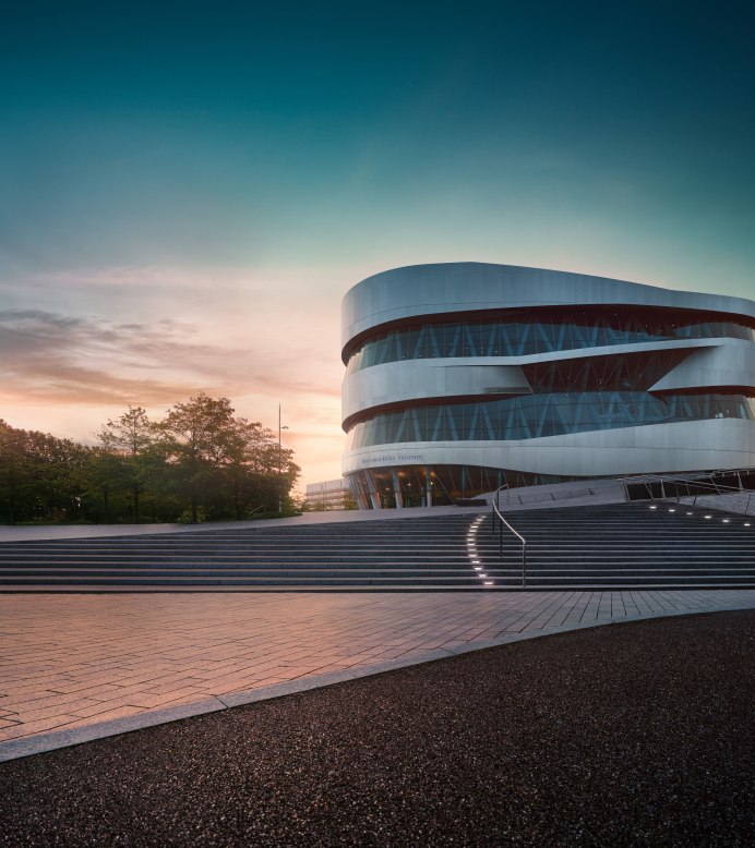 Das Mercedes-Benz Museum in Stuttgart bei Sonnenuntergang. Moderne Architektur mit geschwungenen Linien, umgeben von Bäumen und beleuchteten Stufen., © Mercedes-Benz Heritage GmbH