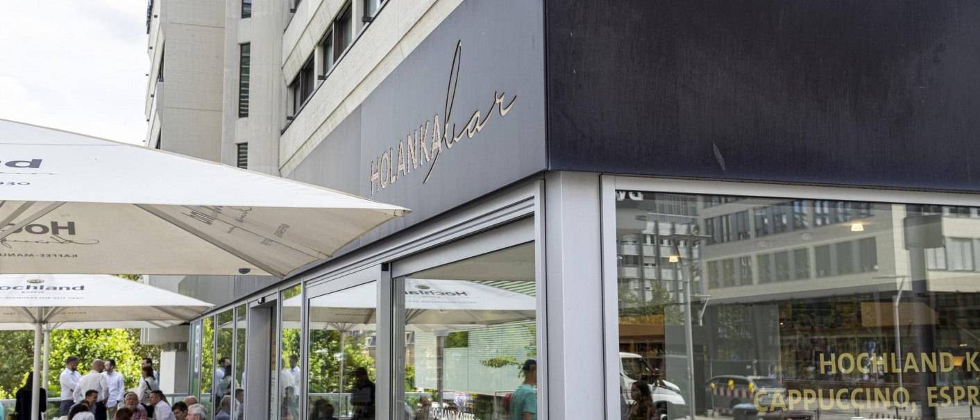 Außenansicht der Holanka Bar mit großen Sonnenschirmen. Menschen sitzen und stehen im Außenbereich. Moderne Gebäude im Hintergrund., © SMG, Sarah Schmid