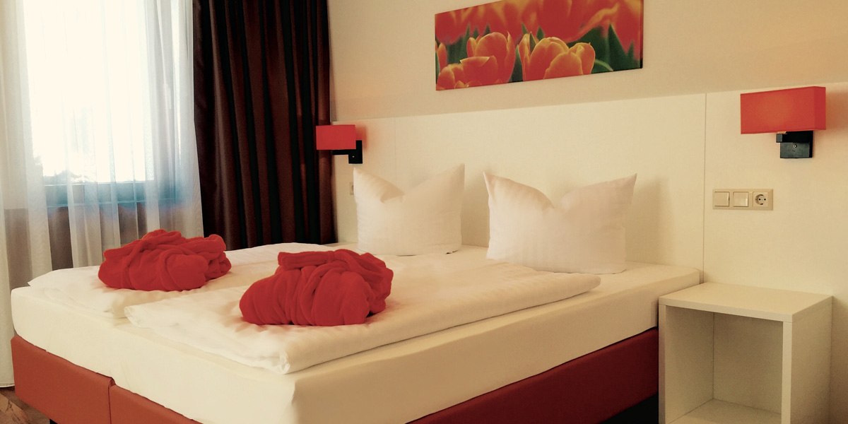 Ein gem&uuml;tliches Hotelzimmer mit Doppelbett, roten Badem&auml;nteln und einem Tulpenbild an der Wand. Die Beleuchtung ist warm und einladend., &copy; TOMAS
