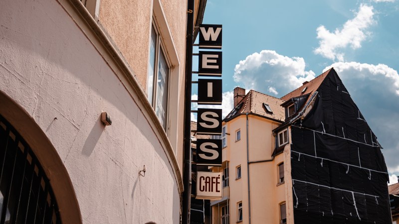 Schild mit 'WEISS Café' an einem Gebäude, daneben ein weiteres Haus mit Gerüst. Blauer Himmel mit Wolken im Hintergrund., © SMG_Sarah Schmid