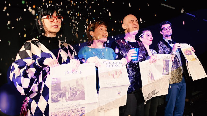 Five people on a stage holding newspapers and surrounded by confetti. They look cheerful and festively dressed., &copy; tri-b&uuml;hne e.V.