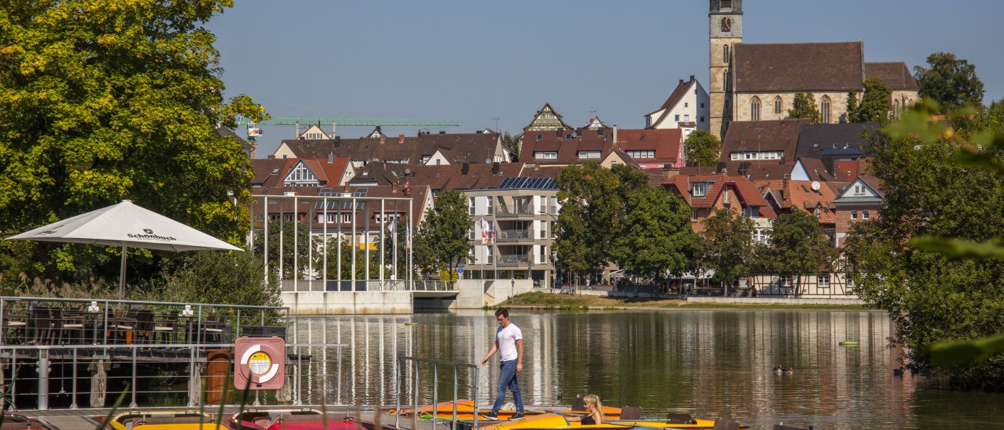 A lake with pedal boats in the foreground, behind it a church and houses in Böblingen. A man walks on the jetty., © Stuttgart-Marketing GmbH, Achim Mende