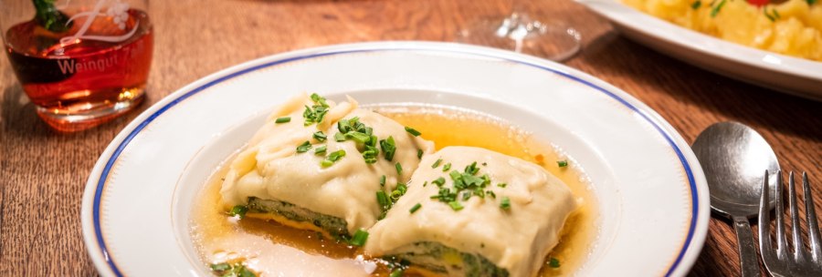 Two Maultaschen in broth on a plate, garnished with chives. Next to it, a glass of red wine and cutlery on a wooden table., © Weingut Zaiß