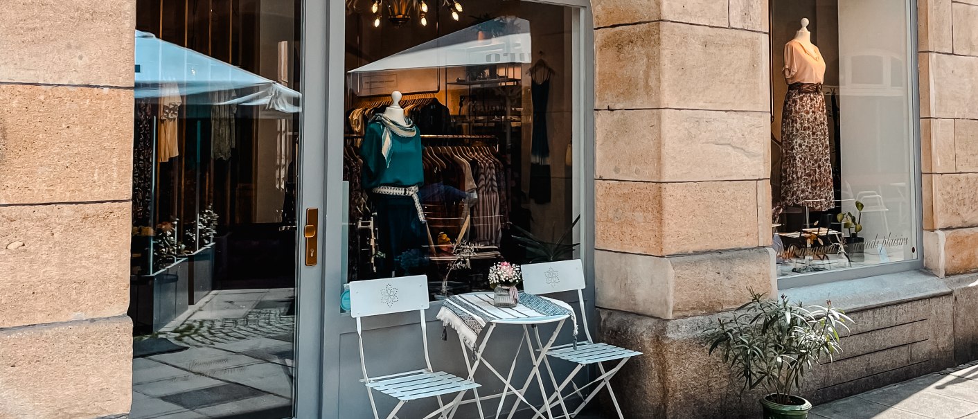 A small store with a display window, decorated with clothes. There are two white chairs and a table on the sidewalk in front of it., © MAGASIN Geißstrasse 15, Stuttgart A small store with a display window, decorated with clothes. There are two white chairs and a table on the sidewalk in front of it., © MAGASIN Geißstrasse 15, Stuttgart