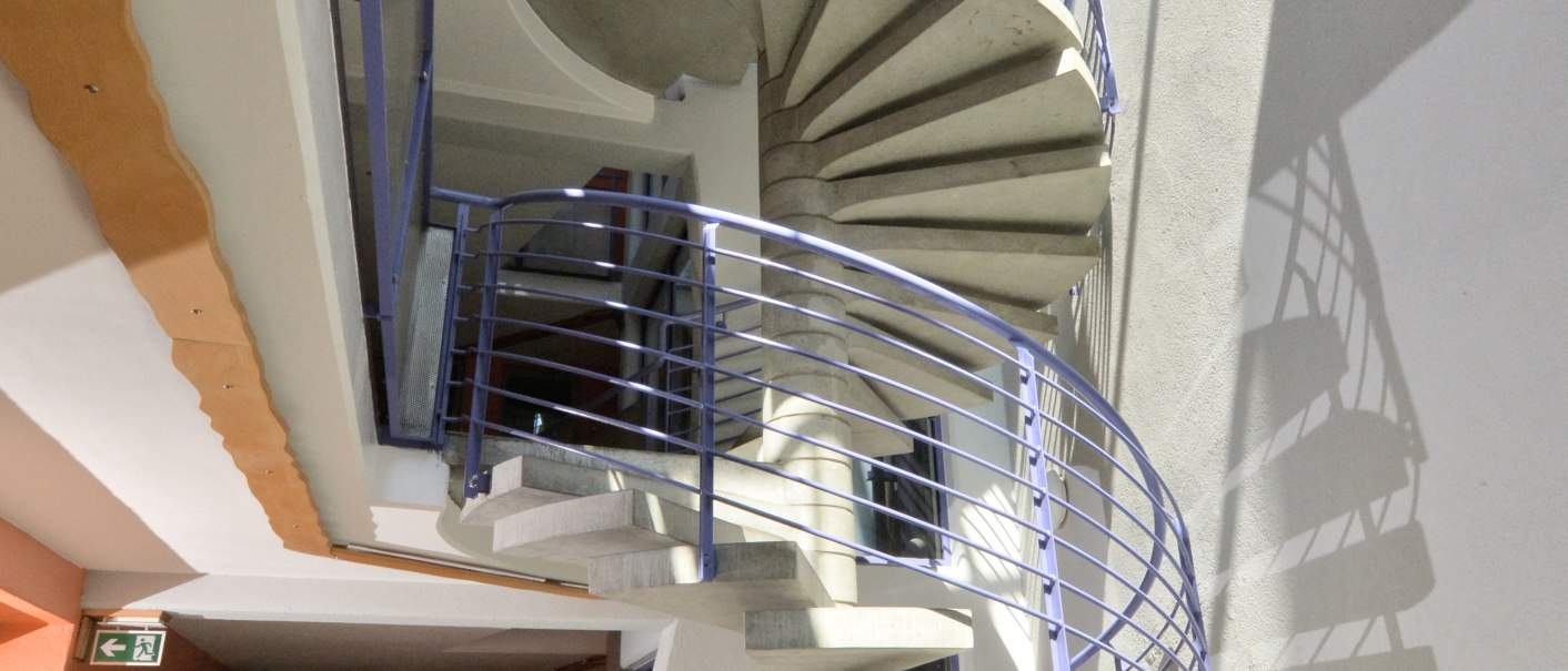 Moderne Wendeltreppe mit blauen Geländern, die sich in einem lichtdurchfluteten Raum mit Glasdach befindet. Unten sind weiße Steine und rote Blumen., © Abalon Hotel Ideal Moderne Wendeltreppe mit blauen Geländern, die sich in einem lichtdurchfluteten Raum mit Glasdach befindet. Unten sind weiße Steine und rote Blumen., © Abalon Hotel Ideal