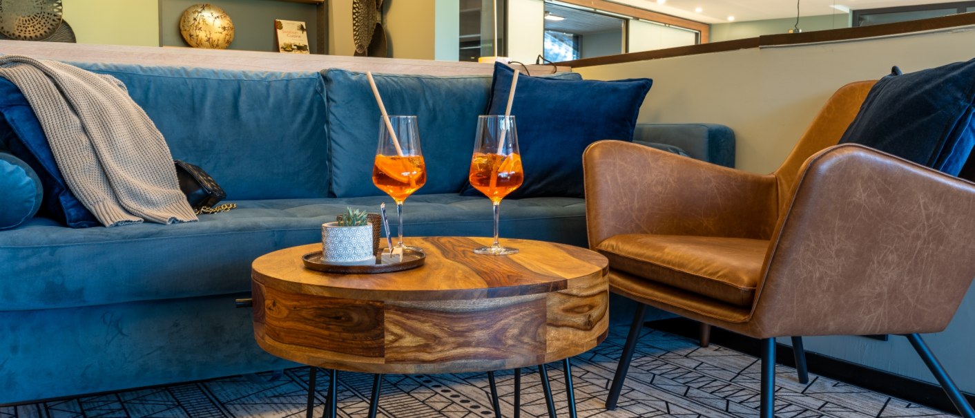 Gem&uuml;tliche Lobby mit blauer Couch, braunem Sessel und Holztisch. Zwei Aperol Spritz Gl&auml;ser stehen auf dem Tisch, im Hintergrund Regale mit Dekoration., &copy; MASEVEN Stuttgart SI-Centrum