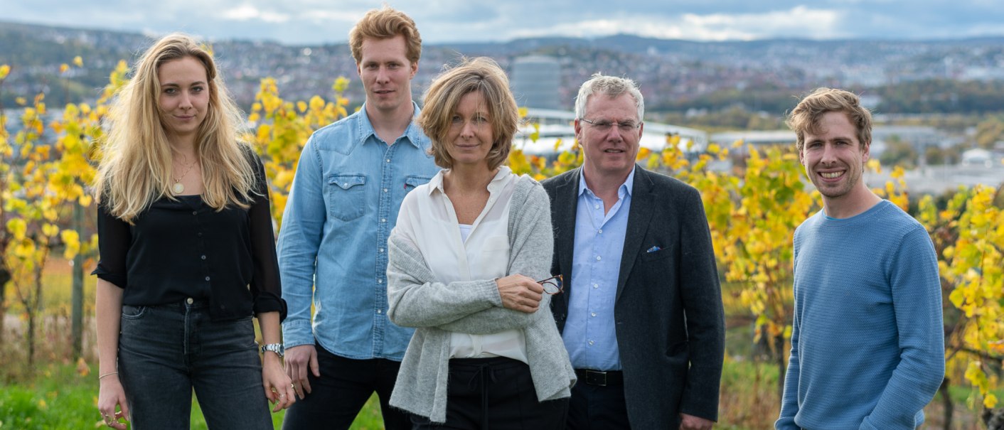 Five people stand smiling in front of an autumnal vineyard. Yellow vine leaves and a cityscape can be seen in the background., © BURKHARDT HELLWIG Five people stand smiling in front of an autumnal vineyard. Yellow vine leaves and a cityscape can be seen in the background., © BURKHARDT HELLWIG