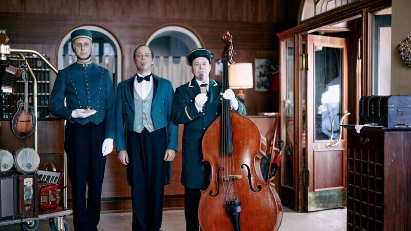 Drei Männer in Hoteluniformen stehen in einer Lobby. Einer hält einen Kontrabass, die anderen tragen Tabletts. Die Szene wirkt wie aus einem alten Hotel., © Theaterhaus Stuttgart e.V. Drei Männer in Hoteluniformen stehen in einer Lobby. Einer hält einen Kontrabass, die anderen tragen Tabletts. Die Szene wirkt wie aus einem alten Hotel., © Theaterhaus Stuttgart e.V.
