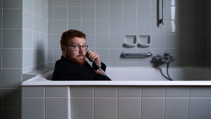 Eine Person mit Brille sitzt in einer Badewanne und hält eine Handbrause. Die Umgebung ist mit grauen Fliesen gestaltet., © Rosenau Kultur e.V.