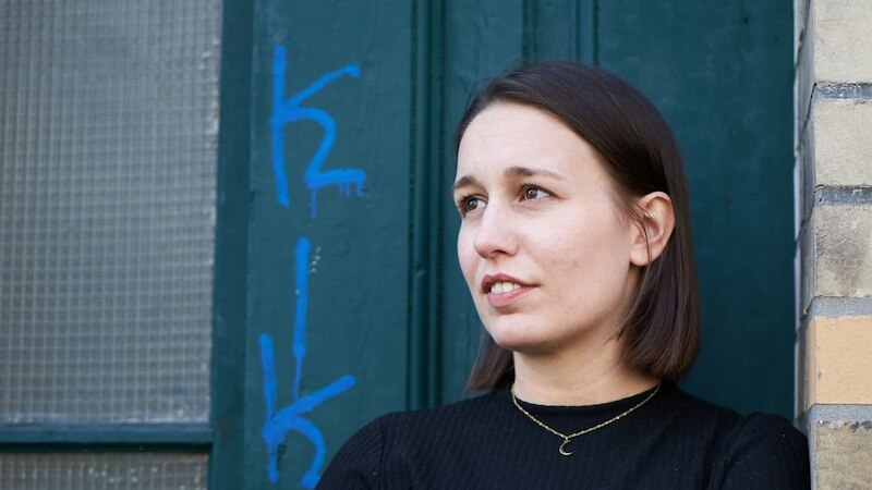 Eine Frau mit braunen Haaren steht vor einer grünen Tür mit blauer Graffiti. Sie trägt ein schwarzes Oberteil und eine goldene Halskette., © Rosenau Kultur e.V. Eine Frau mit braunen Haaren steht vor einer grünen Tür mit blauer Graffiti. Sie trägt ein schwarzes Oberteil und eine goldene Halskette., © Rosenau Kultur e.V.