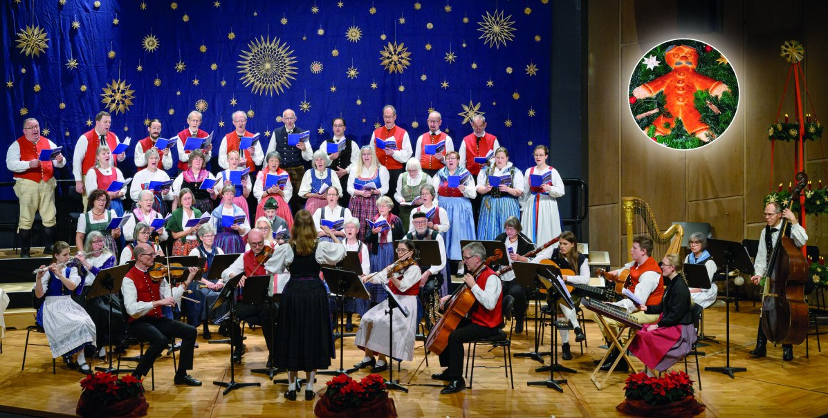Ein Chor und Orchester in traditioneller Kleidung auf einer Bühne. Im Hintergrund weihnachtliche Dekoration mit Sternen und einem Lebkuchenmann., © Stuttgarter Advents-Singen