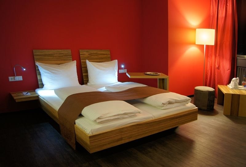 Modernes Hotelzimmer mit roter Wand, Doppelbett, Nachttischlampen und Stehlampe. Elegante Einrichtung mit Holzmöbeln und warmem Licht., © AMH Airport-Messe-Hotel