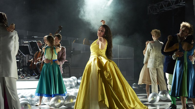 Menschen in eleganter Kleidung tanzen auf einer Bühne. Eine Frau im gelben Kleid steht im Vordergrund. Musiker sind im Hintergrund zu sehen., © Württembergische Staatstheater Stuttgart