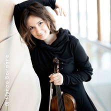 Eine Frau in schwarzer Kleidung lehnt lächelnd an einer Wand und hält eine Geige. Der Hintergrund ist hell und unscharf., © links im Bild
