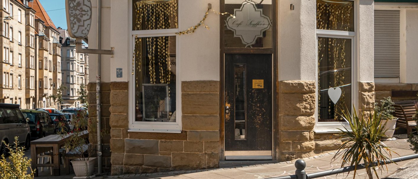 Eingang des Café Seyffer’s an einer Straßenecke, dekoriert mit goldenen Girlanden und Pflanzen. Die Straße ist von Wohnhäusern gesäumt., © SMG, Sarah Schmid Eingang des Café Seyffer’s an einer Straßenecke, dekoriert mit goldenen Girlanden und Pflanzen. Die Straße ist von Wohnhäusern gesäumt., © SMG, Sarah Schmid