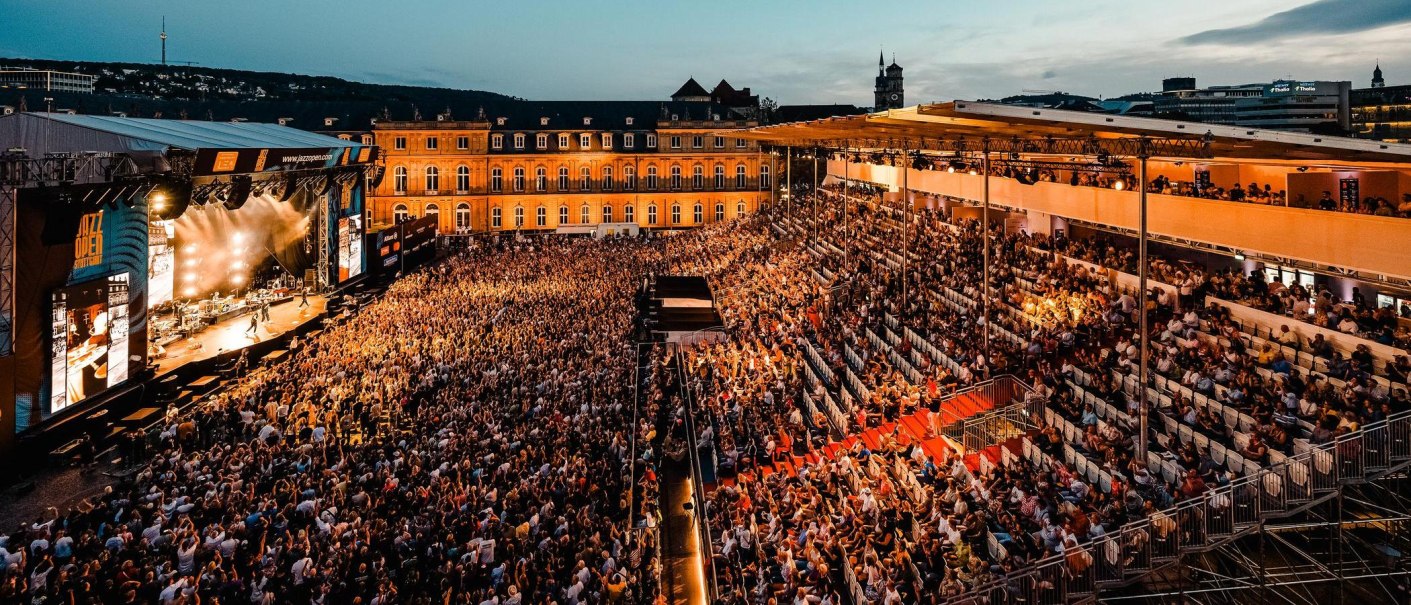 Großes Open-Air-Konzert bei Nacht mit einer Menschenmenge vor einer beleuchteten Bühne. Im Hintergrund sind Gebäude und ein Turm zu sehen., © jazzopen stuttgart, Reiner Pfisterer Großes Open-Air-Konzert bei Nacht mit einer Menschenmenge vor einer beleuchteten Bühne. Im Hintergrund sind Gebäude und ein Turm zu sehen., © jazzopen stuttgart, Reiner Pfisterer