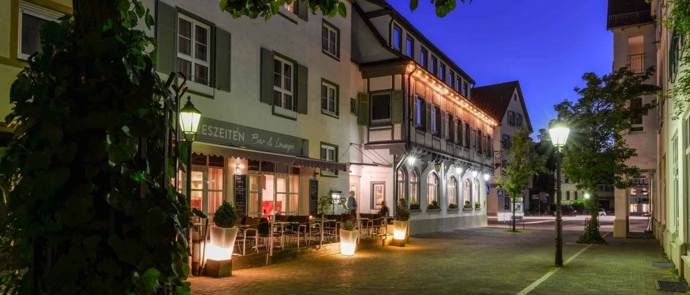 A hotel building lit up at night, surrounded by trees and streetlights. There are tables and chairs in front of the hotel., &copy; Flairhotel Vier Jahreszeiten