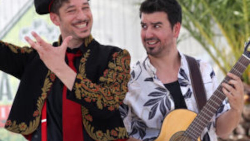 Zwei M&auml;nner, einer in spanischer Tracht mit roter Krawatte, der andere mit Gitarre, stehen unter Palmen und l&auml;cheln fr&ouml;hlich., &copy; Theaterhaus Stuttgart e.V.