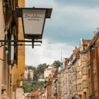 Straßenszene in Stuttgart mit einem Gasthausschild "Das Lehen" und historischen Gebäuden im Hintergrund., © SMG, Sarah Schmid