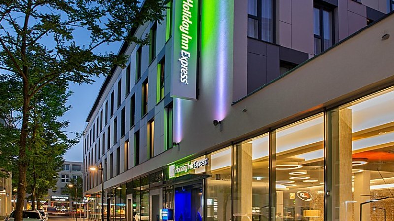 Außenansicht eines modernen Holiday Inn Express Hotels bei Dämmerung, mit beleuchtetem Logo und Glasfassade., © Holiday Inn Express Waiblingen