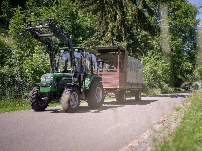 Ein gr&uuml;ner Traktor zieht einen &uuml;berdachten Anh&auml;nger auf einer l&auml;ndlichen Stra&szlig;e, umgeben von &uuml;ppigem Gr&uuml;n und B&auml;umen., &copy; Cool-Tours StattReisen