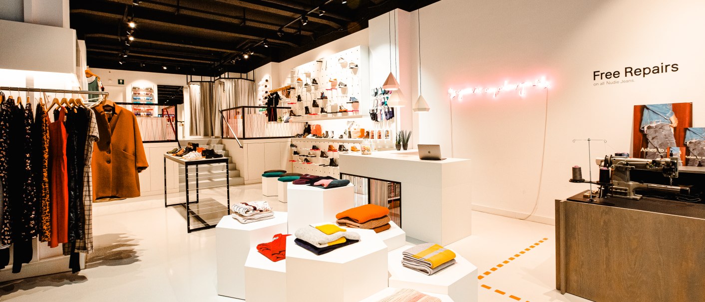 Modernes Bekleidungsgeschäft mit Kleidung auf Ständern, Regalen mit Accessoires und einer Nähmaschine für kostenlose Reparaturen., © Elisa Mauruschat Modernes Bekleidungsgeschäft mit Kleidung auf Ständern, Regalen mit Accessoires und einer Nähmaschine für kostenlose Reparaturen., © Elisa Mauruschat