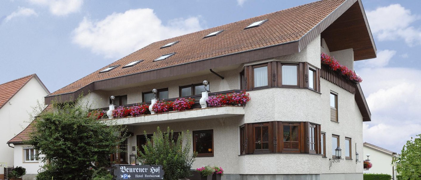 Ein Hotel mit rotem Ziegeldach, Holzbalkonen und blühenden Blumen. Ein Schild zeigt 'Beurener Hof'. Der Himmel ist blau mit wenigen Wolken., © Hotel & Restaurant Beurener Hof Ein Hotel mit rotem Ziegeldach, Holzbalkonen und blühenden Blumen. Ein Schild zeigt 'Beurener Hof'. Der Himmel ist blau mit wenigen Wolken., © Hotel & Restaurant Beurener Hof