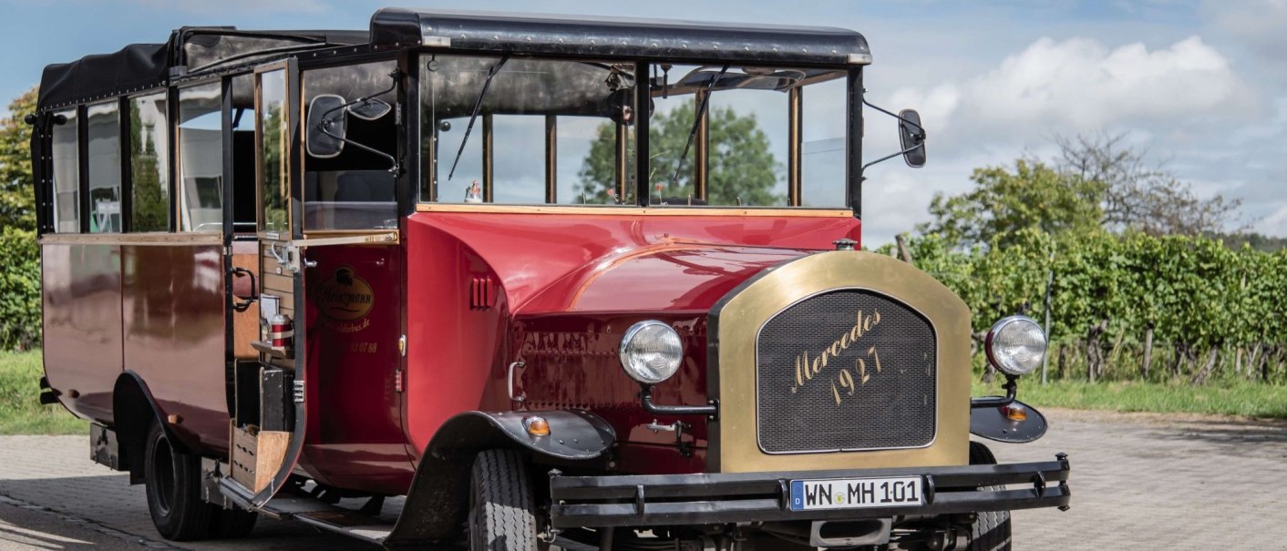 Roter Cabrio-Oldtimerbus mit Mercedes-Logo, Baujahr 1927, auf gepflastertem Weg vor grüner Landschaft., © Stuttgart-Marketing GmbH, trickytine Roter Cabrio-Oldtimerbus mit Mercedes-Logo, Baujahr 1927, auf gepflastertem Weg vor grüner Landschaft., © Stuttgart-Marketing GmbH, trickytine