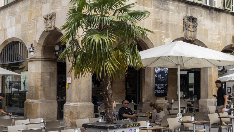 Caf&eacute; with tables and chairs under parasols in front of a historic building with arcades. Palm trees and people in the outdoor area., &copy; SMG, Sarah Schmid