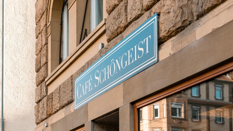 Das Caf&eacute; Sch&ouml;ngeist zeigt ein blaues Schild an einer historischen Steinwand. Im Fenster spiegelt sich die umliegende Architektur wider., &copy; SMG Stuttgart Marketing GmbH - Sarah Schmid