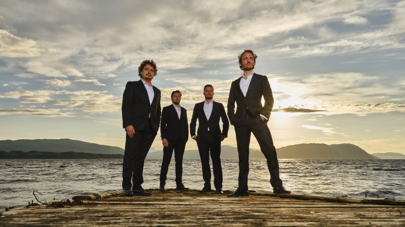 Vier Männer in Anzügen posieren auf einem Steg vor einer malerischen Kulisse mit Wasser und Bergen im Sonnenuntergang., © ECHELON Vier Männer in Anzügen posieren auf einem Steg vor einer malerischen Kulisse mit Wasser und Bergen im Sonnenuntergang., © ECHELON