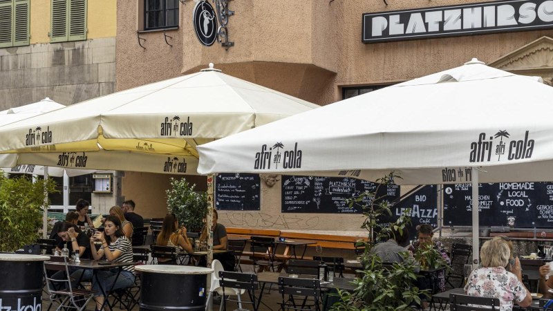 Außenbereich eines Cafés namens Platzhirsch mit Sonnenschirmen und Gästen. Menschen sitzen an Tischen und genießen Getränke., © SMG, Sarah Schmid