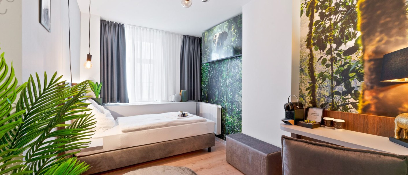 Ein modernes Einzelzimmer mit Dschungelmotiv an der Wand, einem Bett, Schreibtisch und Pflanzen. Helle Beleuchtung und stilvolle Einrichtung., &copy; JUNGLE N&deg;5 GmbH