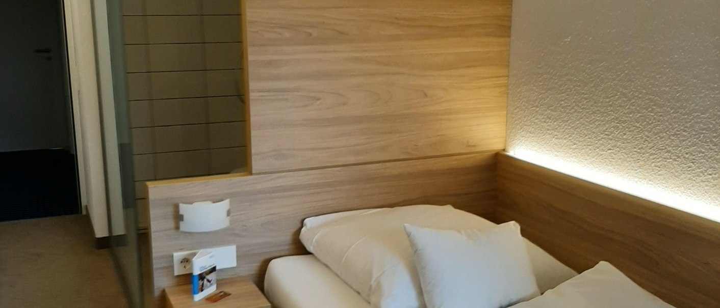 Ein gemütliches Einzelzimmer mit einem Bett, einem kleinen Nachttisch und einer offenen Dusche im Hintergrund., © Hotel am Schlossberg Herrenberg