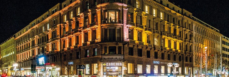 A large, illuminated building at night with stores on the first floor and people on the street., © Schauspielbühnen in Stuttgart A large, illuminated building at night with stores on the first floor and people on the street., © Schauspielbühnen in Stuttgart