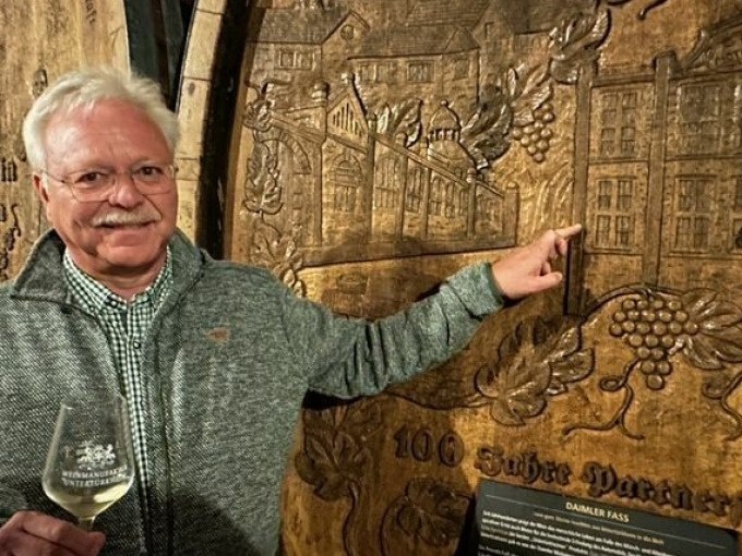 Ein Mann zeigt auf ein kunstvoll geschnitztes Weinfass in einem Weinkeller. Er hält ein Weinglas und lächelt in die Kamera., © Weingenuss Stuttgart