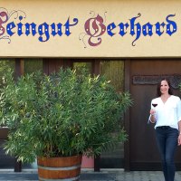 Eine Frau steht vor dem Weingut Gerhard Zaiß, hält ein Glas Rotwein und lächelt. Das Gebäude hat eine dekorative Beschriftung und Pflanzen im Vordergrund., © Simone Mack Eine Frau steht vor dem Weingut Gerhard Zaiß, hält ein Glas Rotwein und lächelt. Das Gebäude hat eine dekorative Beschriftung und Pflanzen im Vordergrund., © Simone Mack