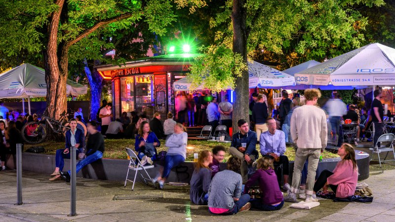 Belebter Außenbereich mit Menschen unter beleuchteten Schirmen und Bäumen. Einige sitzen auf Stühlen, andere auf dem Boden. Bunte Lichter im Hintergrund., © TMBW, Gregor Lengler