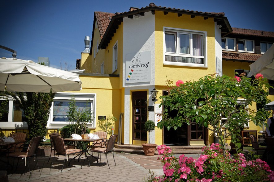 Gelbes Gebäude mit Terrasse, Tischen und Stühlen. Sonnenschirme und blühende Pflanzen im Vordergrund. Schild mit der Aufschrift 'römerhof' an der Fassade., © Römerhof Hotelbetriebs GmbH