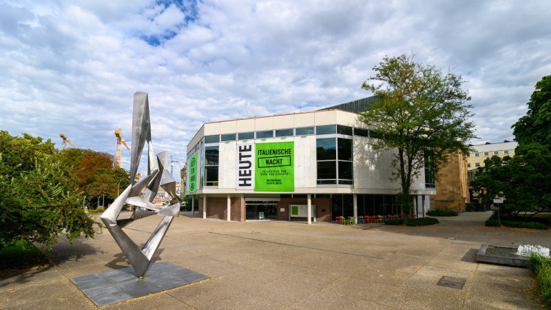 Moderne Skulptur vor dem Schauspielhaus mit großen Fenstern und Plakaten für 'Italienische Nacht'. Wolkenbedeckter Himmel, Bäume und Baukräne im Hintergrund., © Björn Klein