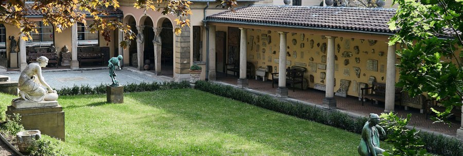 A quiet courtyard with statues and antique relics on the walls. The lawn is well-tended and trees provide shade. Arcades frame the courtyard., &copy; &copy; St&auml;dtisches Lapidarium, Foto Julia Ochs