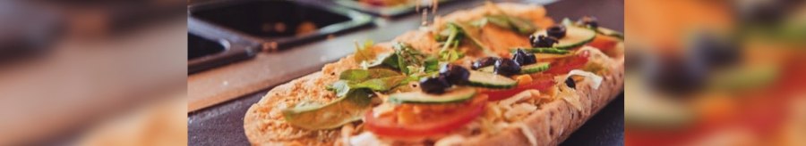 Ein Sandwich wird mit frischem Gemüse und Oliven belegt. Eine Hand streut Gewürze darüber. Im Hintergrund sind Zutaten in Behältern zu sehen., © FreshSub