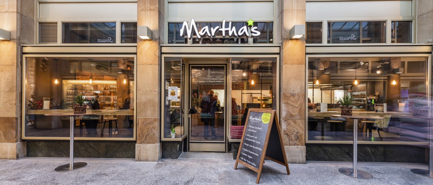 Eingang des Cafés Martha's mit großen Fenstern, Tischen und einer Speisekarte im Freien. Innen sind Lichter und Dekorationen sichtbar., © Matha's, Stuttgart