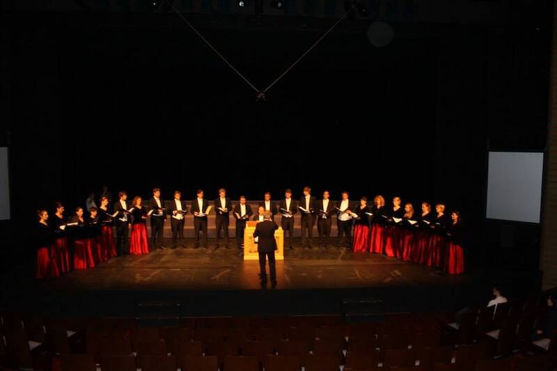 Ein Chor in formeller Kleidung singt auf einer Bühne, dirigiert von einer Person in der Mitte. Die Bühne ist dunkel, der Fokus liegt auf den Sängern., © Musik Podium Stuttgart e.V. Ein Chor in formeller Kleidung singt auf einer Bühne, dirigiert von einer Person in der Mitte. Die Bühne ist dunkel, der Fokus liegt auf den Sängern., © Musik Podium Stuttgart e.V.