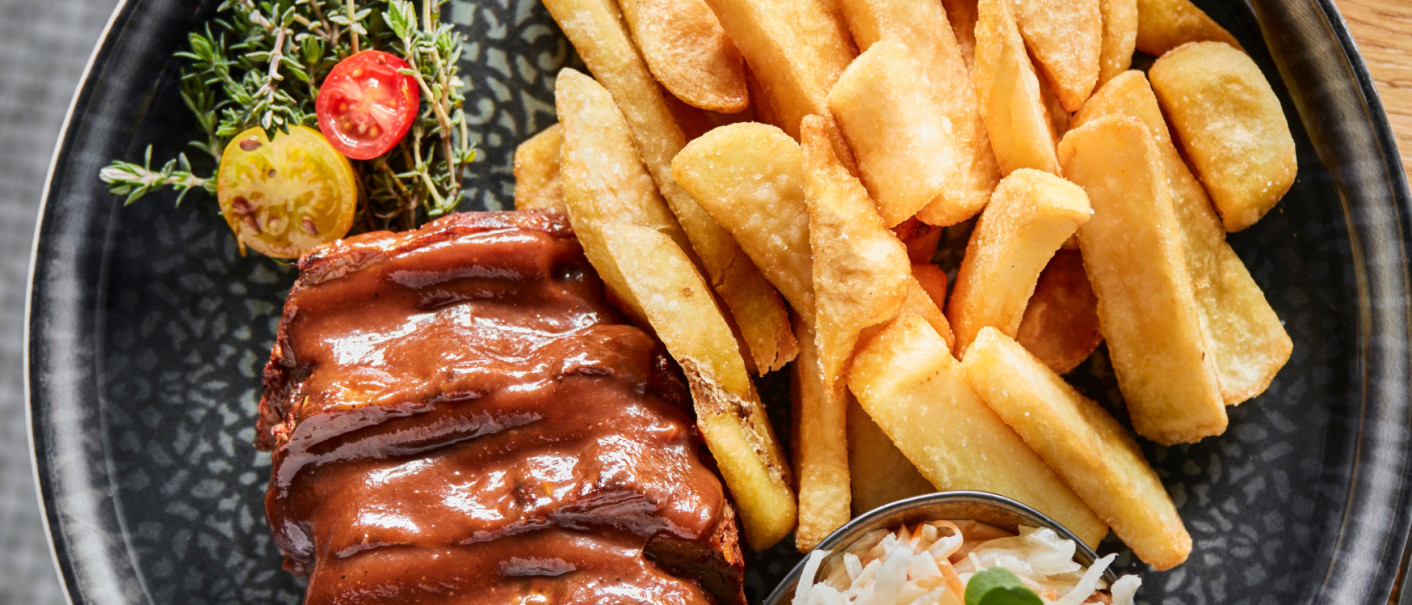 A plate of juicy ribs in sauce, crispy chips and a portion of coleslaw, garnished with herbs and tomatoes., © MAREDO