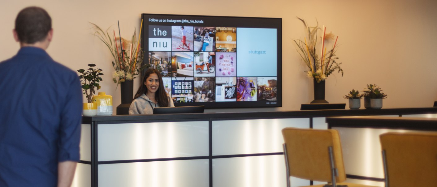 A woman is standing at a hotel front desk, smiling. In the background, there is a screen displaying images and text. Decorations and plants adorn the counter., © the niu Form - Novum Hospitality A woman is standing at a hotel front desk, smiling. In the background, there is a screen displaying images and text. Decorations and plants adorn the counter., © the niu Form - Novum Hospitality