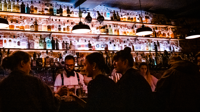 Eine stimmungsvolle Bar mit beleuchteten Flaschenregalen. Menschen unterhalten sich, w&auml;hrend ein Barkeeper Getr&auml;nke zubereitet., &copy; Stuttgart-Marketing GmbH, Sarah Schmid
