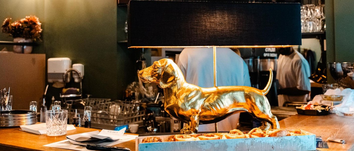 Eine goldene Dackel-Lampe steht auf einer Theke in einem Café. Im Hintergrund sind Gläser und eine Person zu sehen., © Stuttgart Marketing GmbH - Sarah Schmid