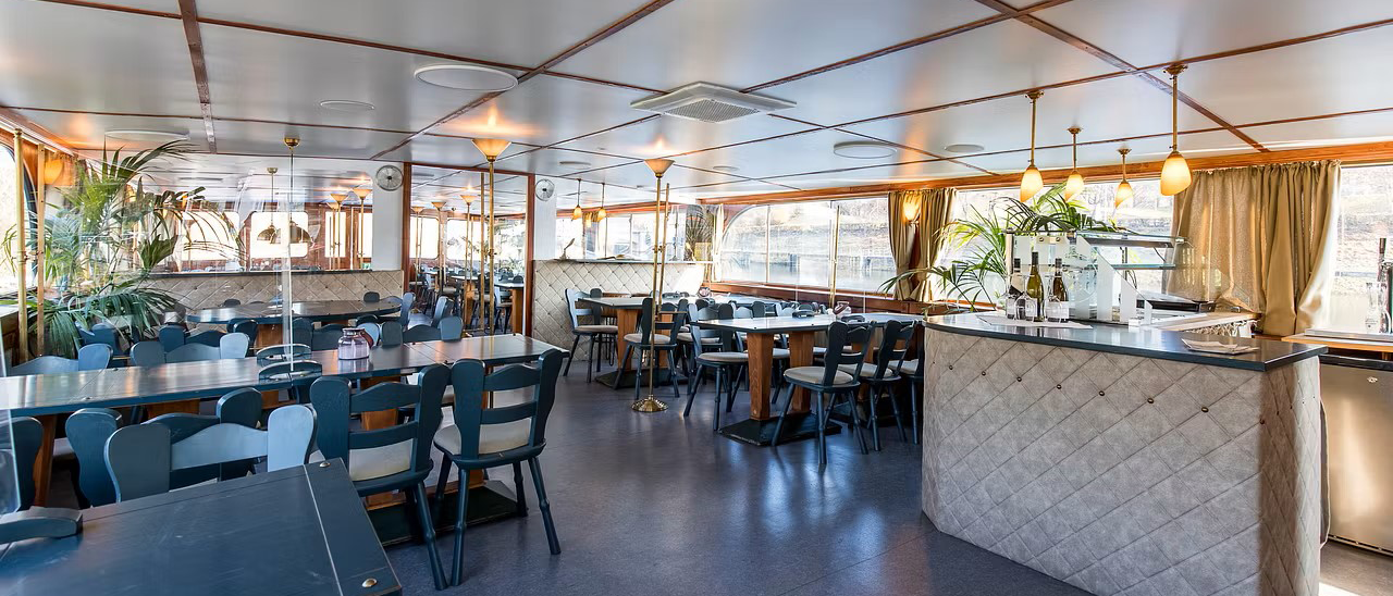 Ein elegantes Schiffsrestaurant mit gedeckten Tischen, blauen St&uuml;hlen und einer Bar. Gro&szlig;e Fenster bieten Ausblick auf das Wasser., &copy; NeckarFlair MS Wilhelma Betriebsgesellschaft mbH